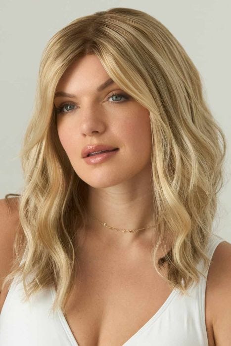 Woman in a just past the shoulders light ash brown wavy wig with long side-swept bangs and dark roots