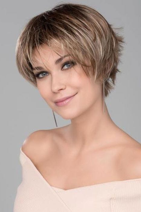 Woman in a long blonde pixie wig with dark roots and short spiky hair in back